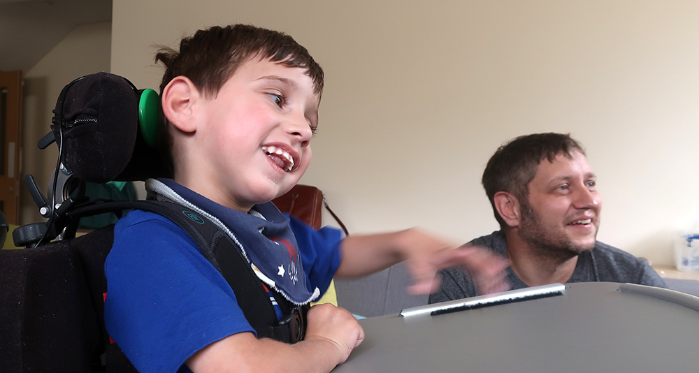 Young boy using adapted gaming controller, smiling man in background