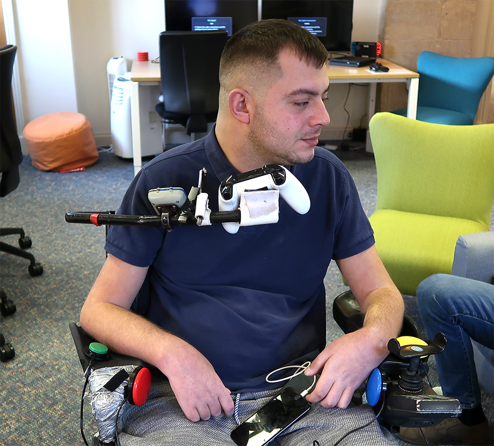 Man in wheelchair with games controller on mounting arm and button switches near his hands