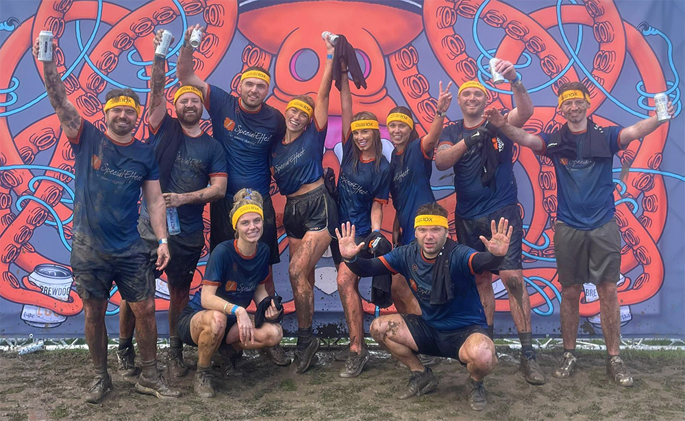 Ten smiling and very muddy runners celebrate completion in front of a large event banner