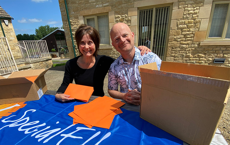 Smiling woman and man seated at outdoor table with boxes of envelopes