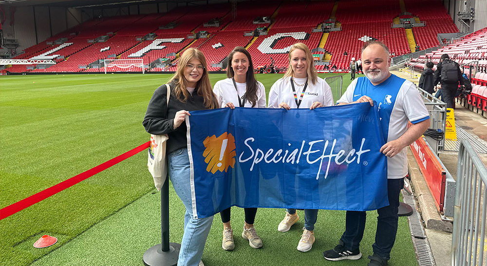 Three women and a man holding a SpecialEffect flag inside a football stadium