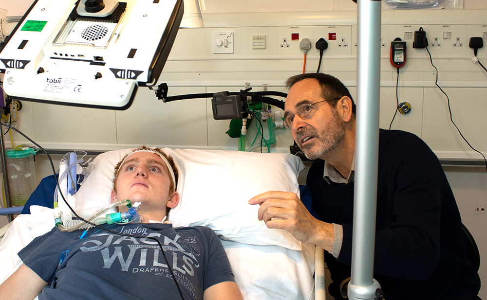 Man in hospital bed looking up at a computer, Mick Donegan watches from bedside