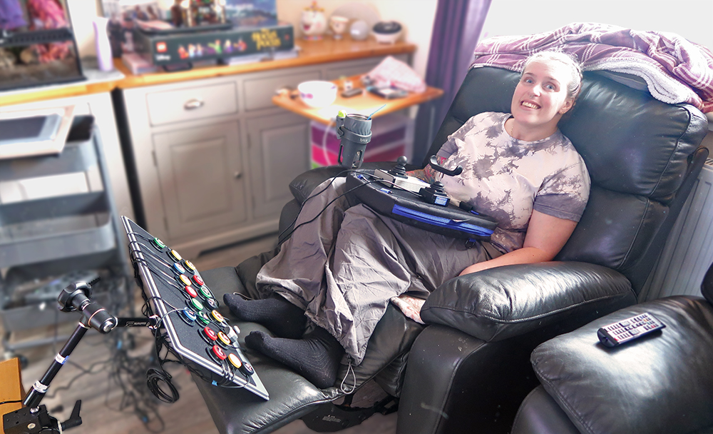 Smiling woman reclining on a sofa seat, with two joystick on the lap and 18 switches on a board by her feet