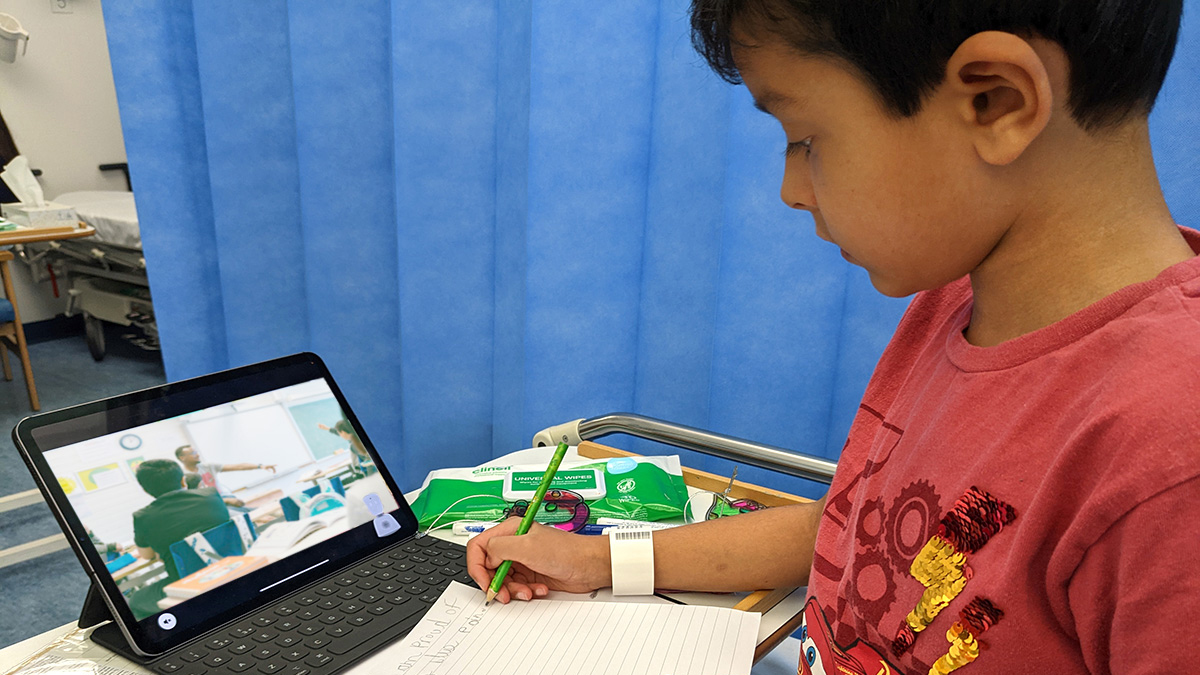 A young boy in hospital writing and looking at the screen of a small tablet computer.