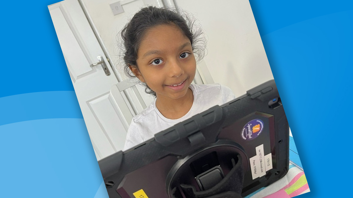 A smiling young girl at home looks over the top of an iPad screen.