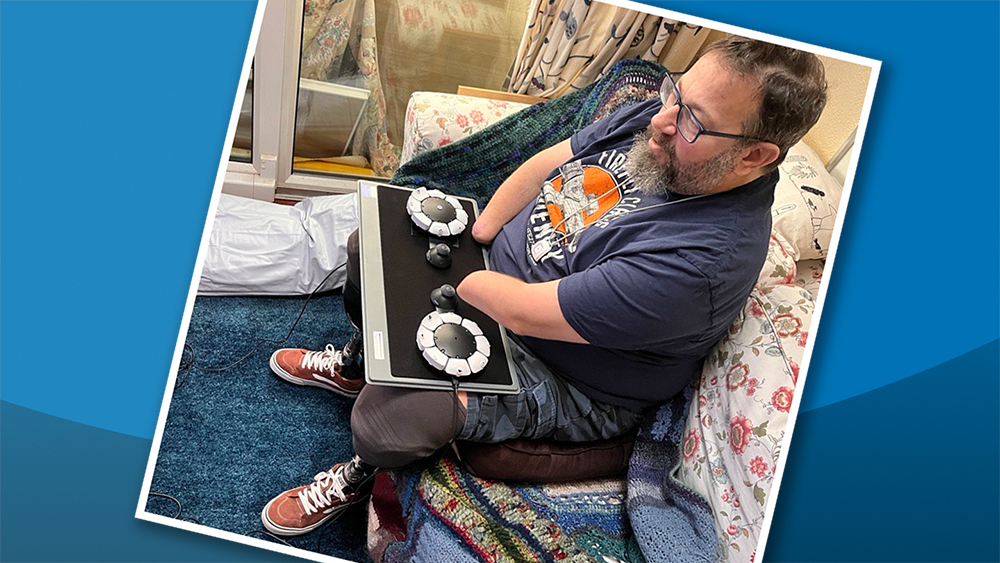 Seated man with beard operating two circular accessible games controllers on a laptray with both part-amputated arms 