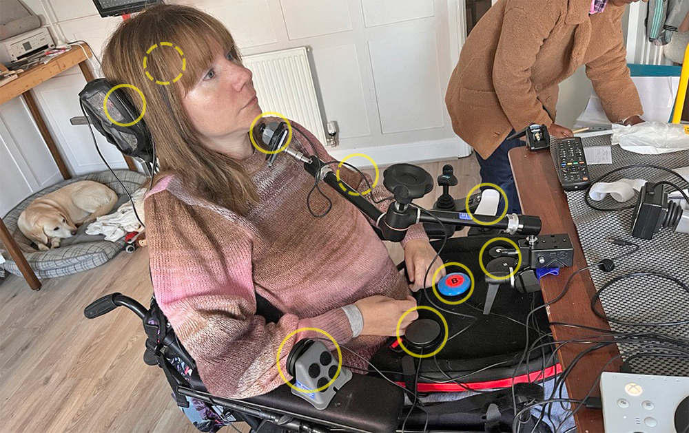 A woman sits in wheelchair with a selection of accessibility switches and joysticks placed around her head, chin, arms and lap tray