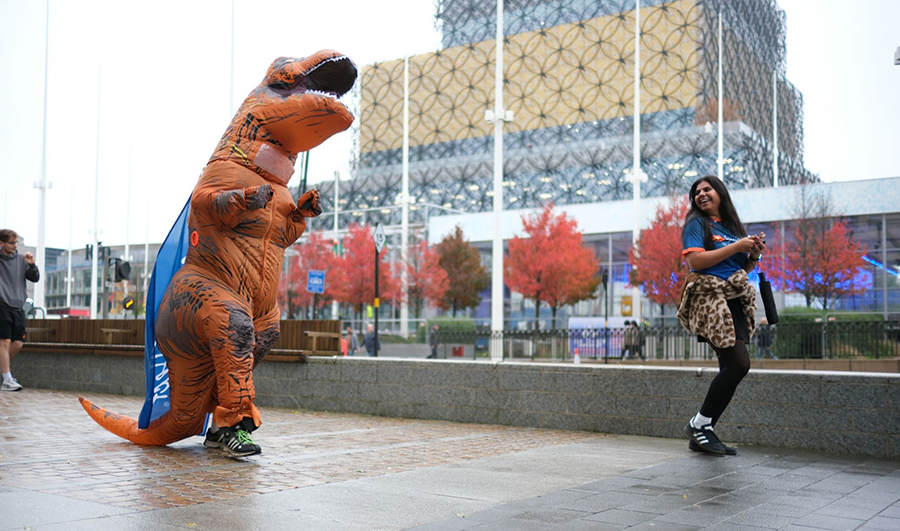 Runner in a T-rex costume on a pavement following a laughing runner in a SpecialEffect T shirt