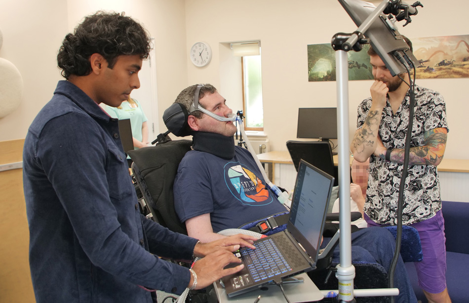 Man in wheelchair looks at screen, therapist types on laptop, another looks on.