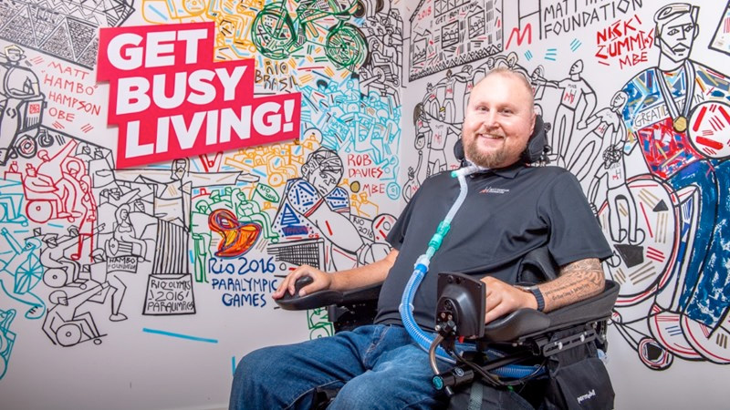 Smiling man in powered wheelchair next to wall with the phrase 'Get Busy Living'