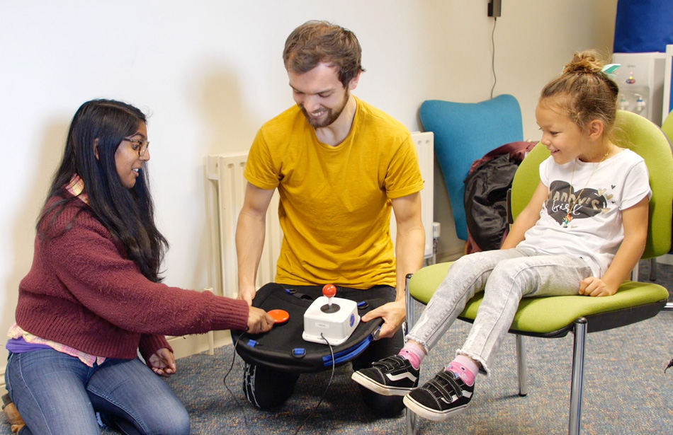 Two therapists prepare an adapted gaming setup for a seated young girl.