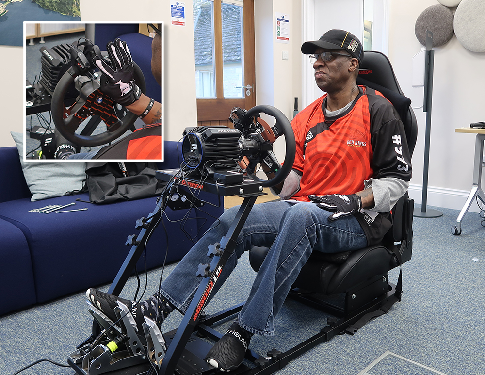 Man sits in a full video game racing rig. He holds a steering wheel with one hand.