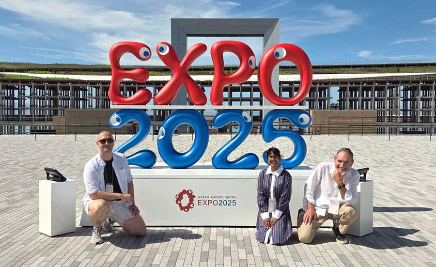 Three people crouch in front of a large sign saying Expo 2025