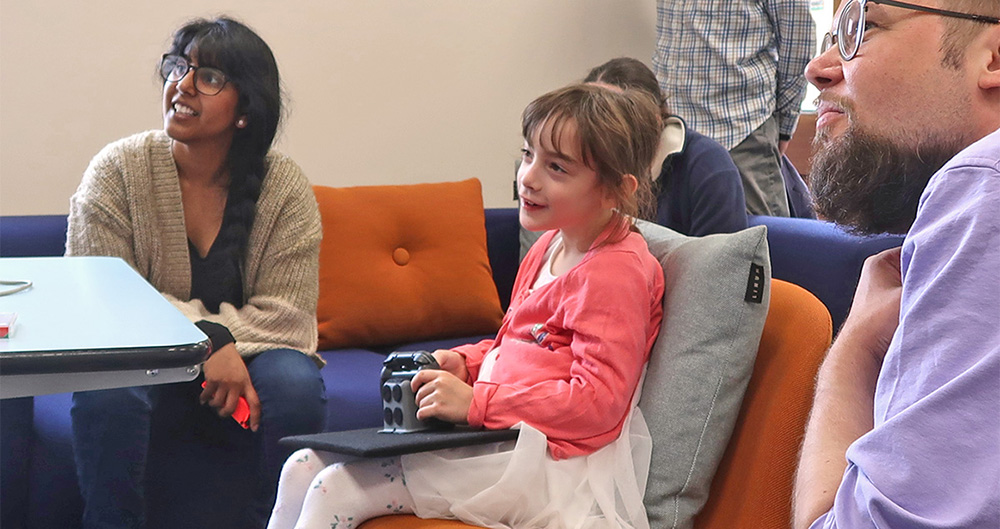 Seated young girl using a games controller on her lap. A smiling woman looks on.